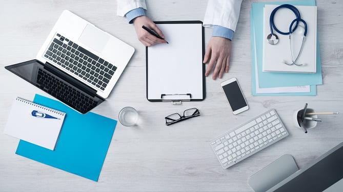 mesa com computador, estetoscópio, bloco de folhas e óculos de médico em quarentena